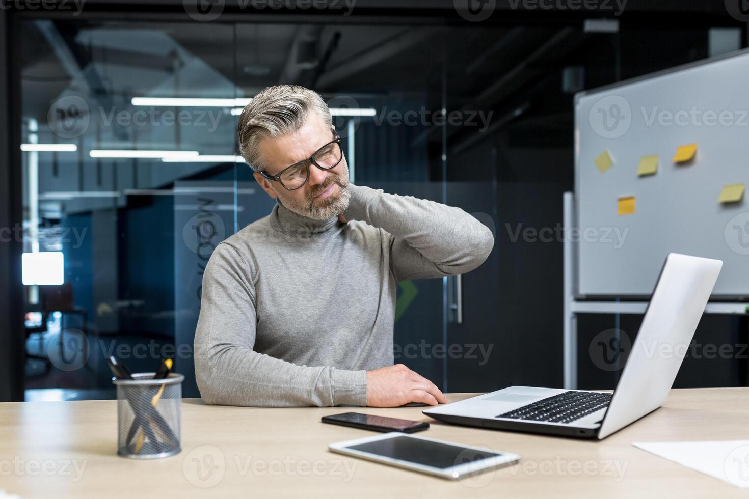Senior mature businessman working inside office building, gray haired man sick and tired massaging neck with hand, boss having severe pain in neck and back. photo