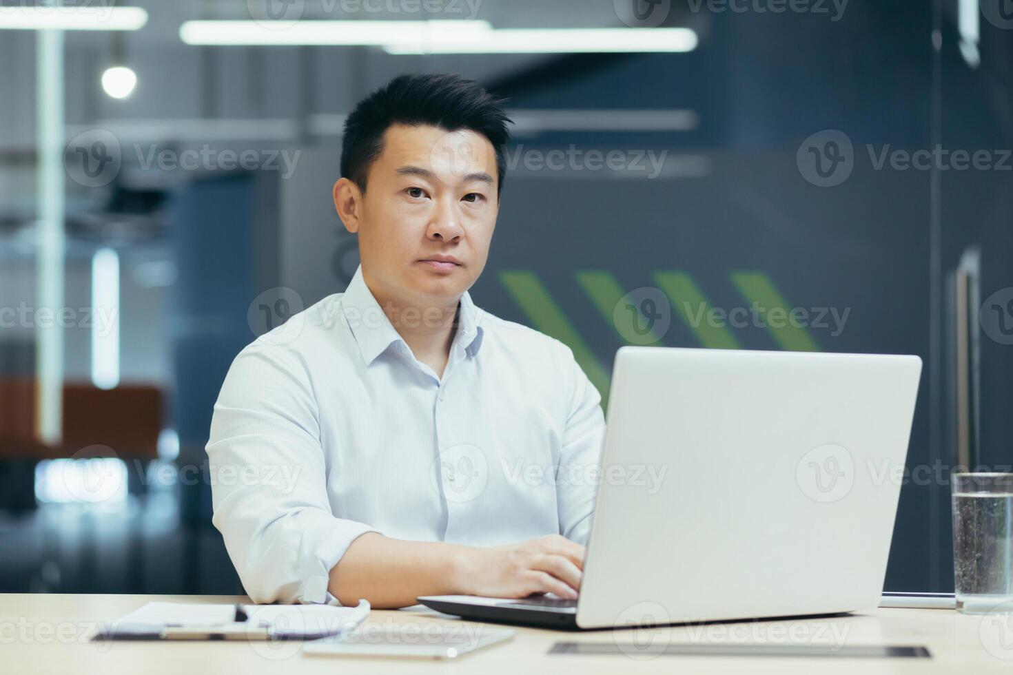 Portrait of successful and serious Asian businessman, man working in modern office typing on laptop, looking at camera. photo