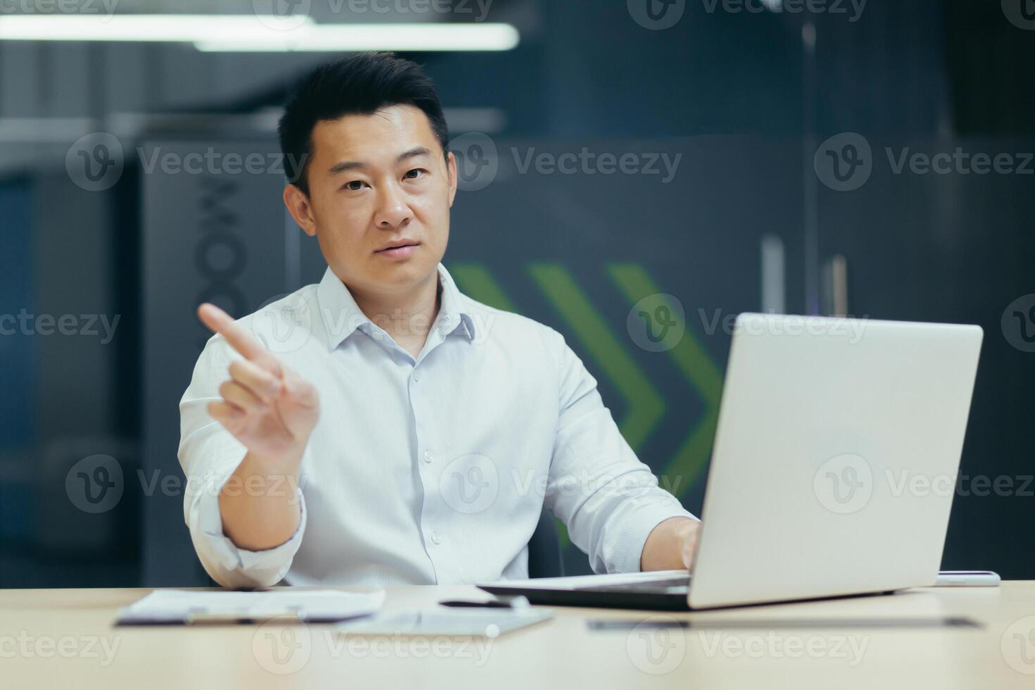 Portrait of a young Asian man, confused, shows with his hands that it is not allowed, did not understand, looks at the camera. Sitting at a desk in the office, working with a laptop online photo