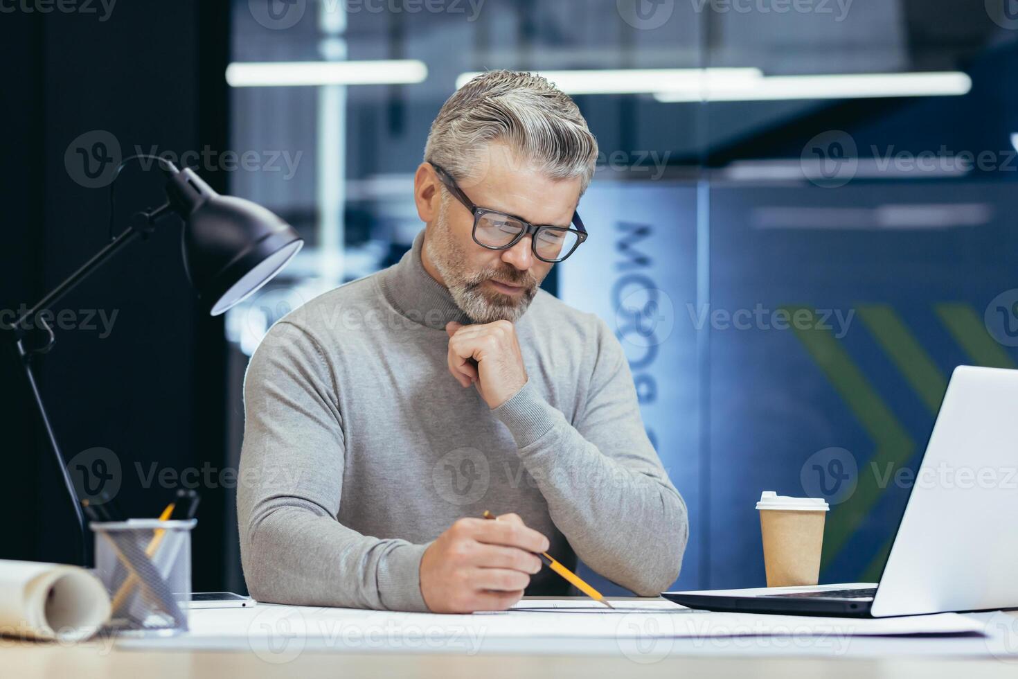 Close-up photo. Concentrated senior gray-haired man architect, engineer, designer sitting in office at table with laptop, working with drawings.plans. Makes notes, paperwork. photo