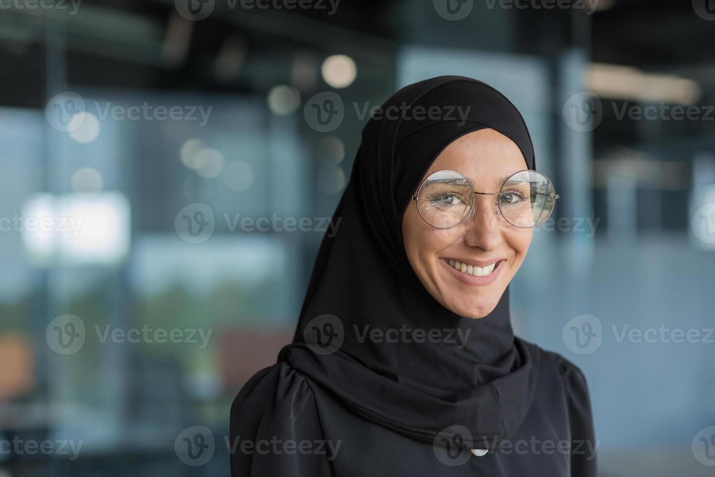 Close-up portrait of Muslim businesswoman in hijab smiling and looking at camera, female worker in glasses working inside modern office building. photo