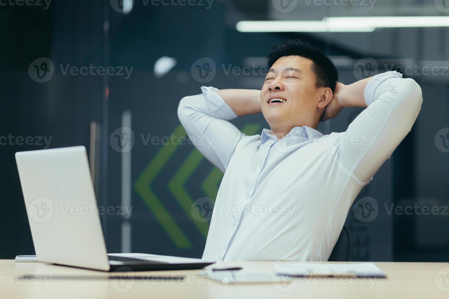 A young handsome Asian man, a businessman sits in the office at the desk. He holds his hands behind his head, closes his eyes, and smiles. Satisfied with his work, rests during working hours. photo