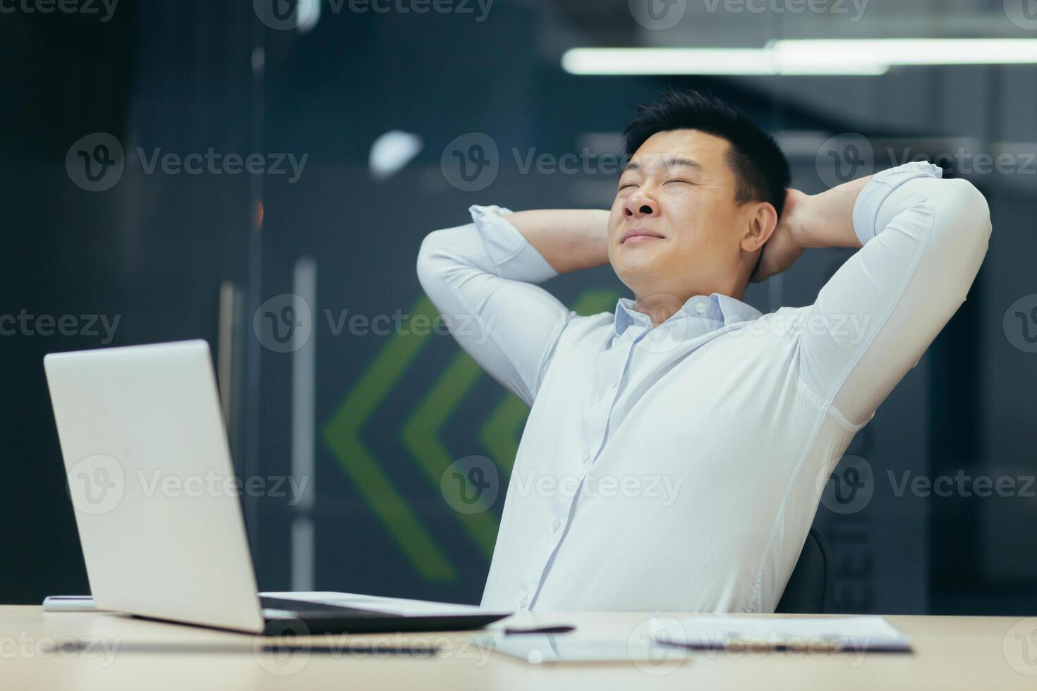 Young Asian male businessman sitting in the office at the desk and resting during the working day. He closed his eyes, threw his hands behind his head. relaxes. photo