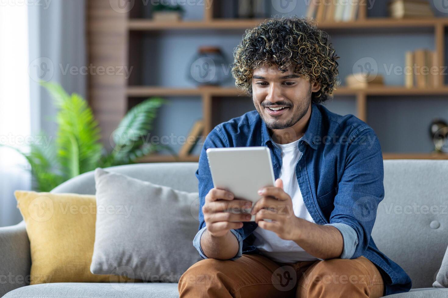 Young man sitting on sofa in living room at home with tablet computer in hands, smiling happily, using online app, browsing social media photo