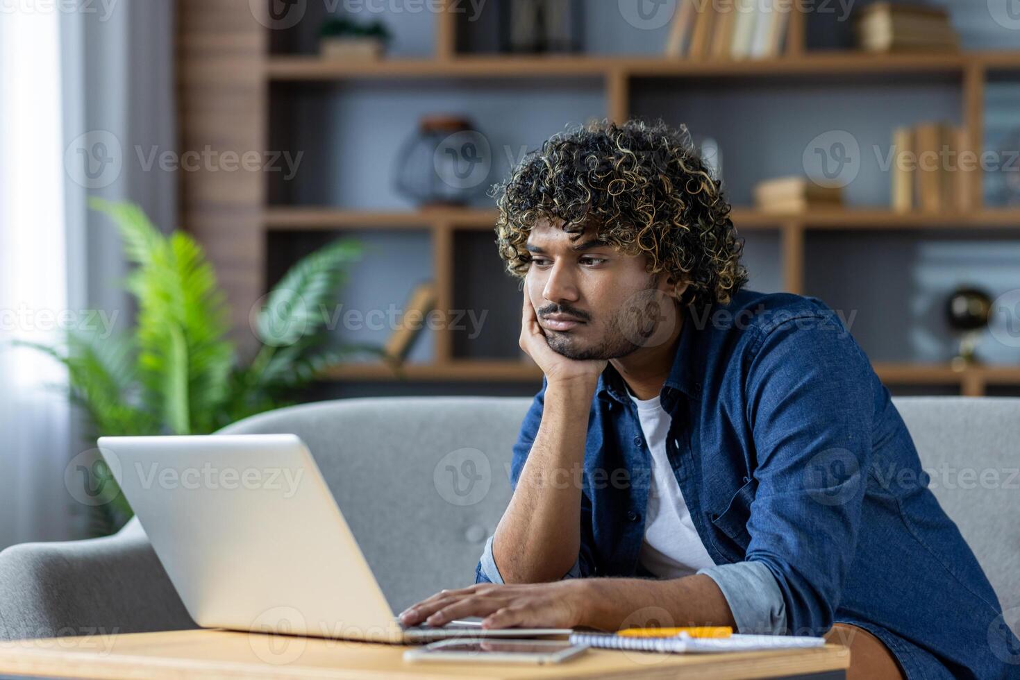 Young man looks bored while working on laptop from home photo