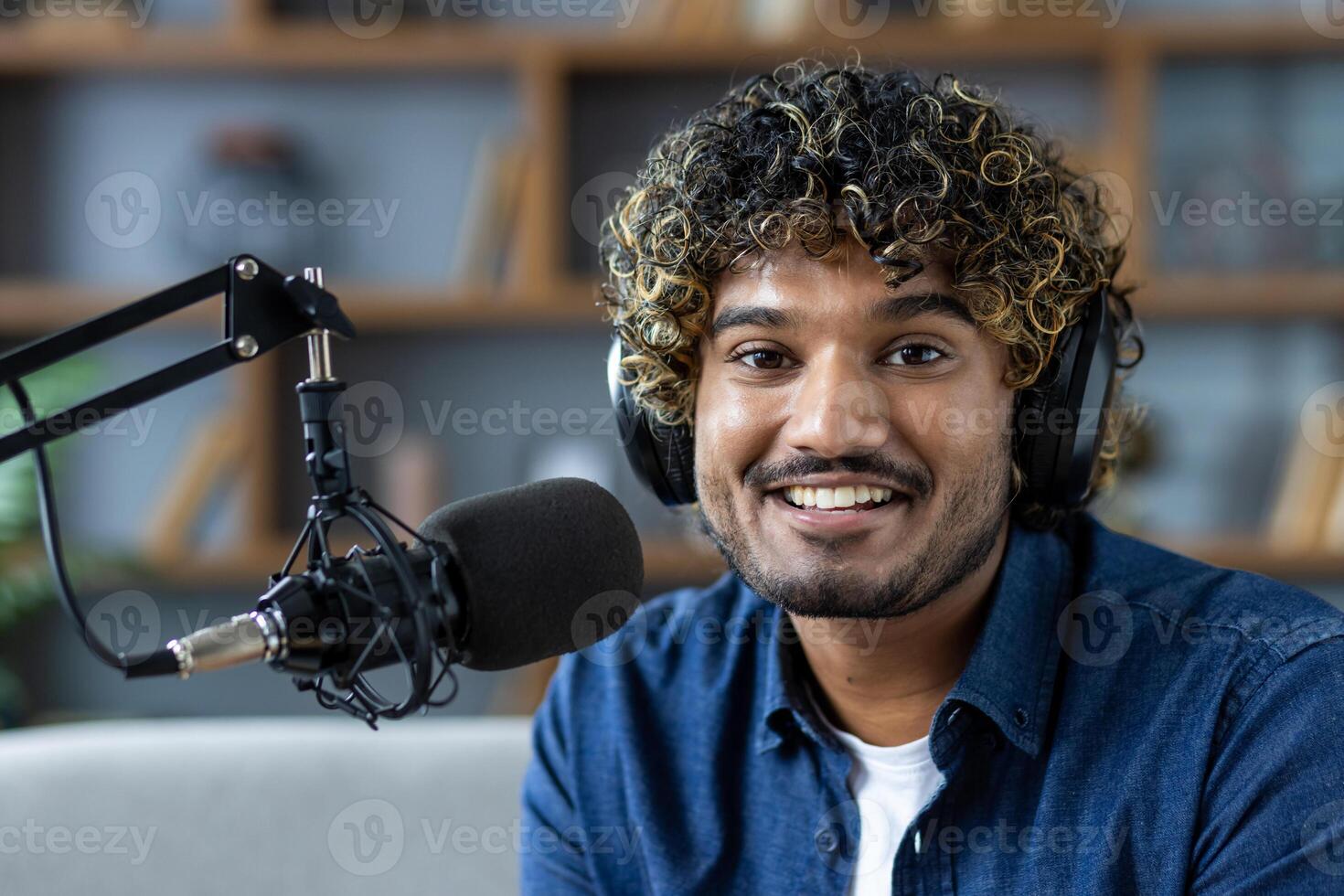 Close-up portrait blogger in headphones and with a professional microphone. The man smiling and looking at camera sitting on sofa in living room at home, recording podcast, conducting online stream photo