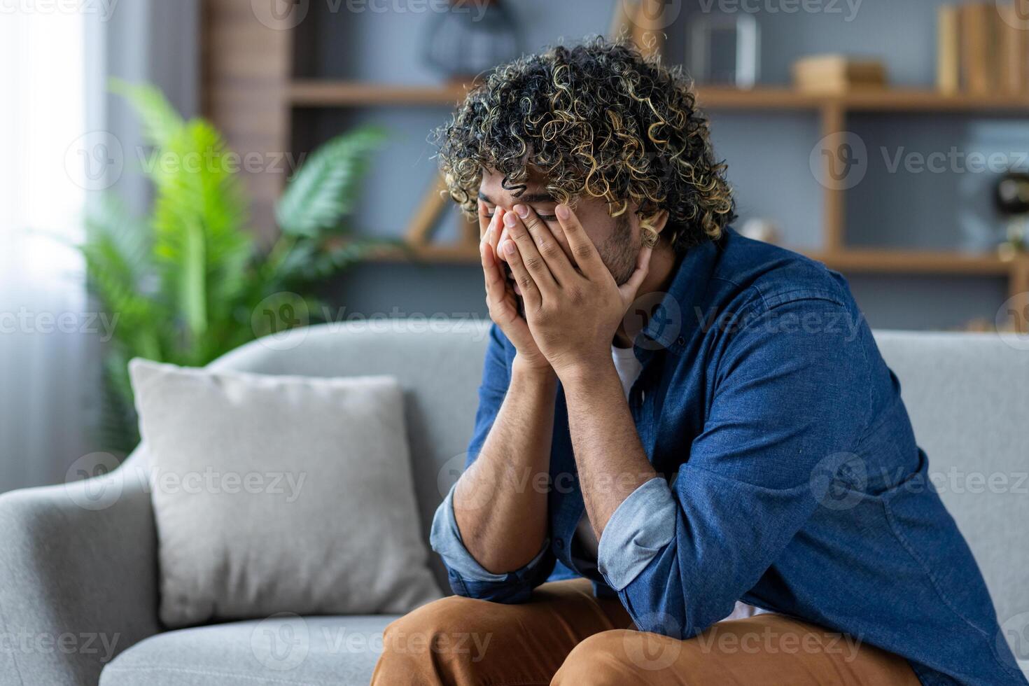 Desperate and depressed man sitting on sofa in living room at home, burying face in hands, feeling sad and disappointed photo