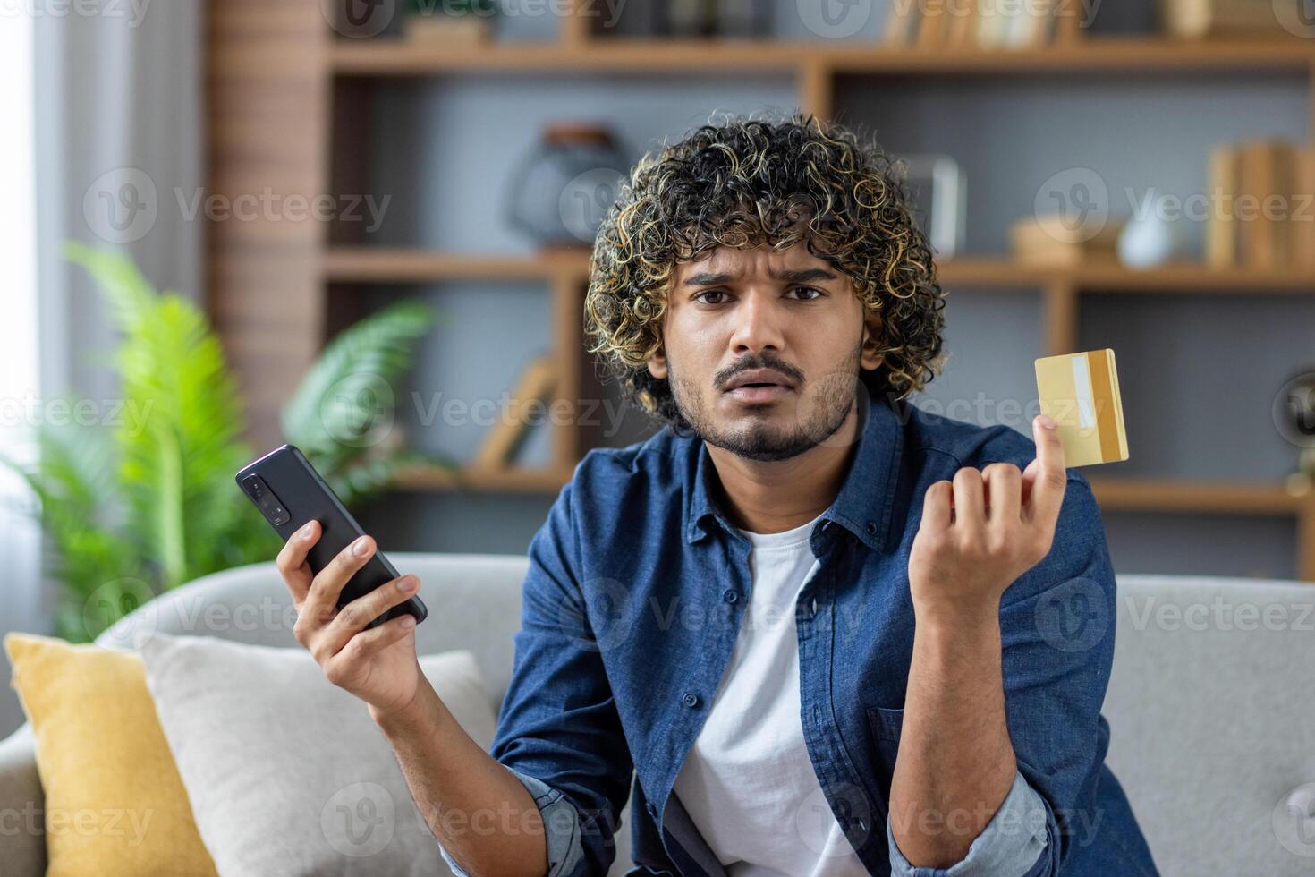 Disappointed and deceived man looking at camera, young freelancer sitting at home on sofa in living room, holding bank credit card and phone, received account blocking, and money transfer error photo