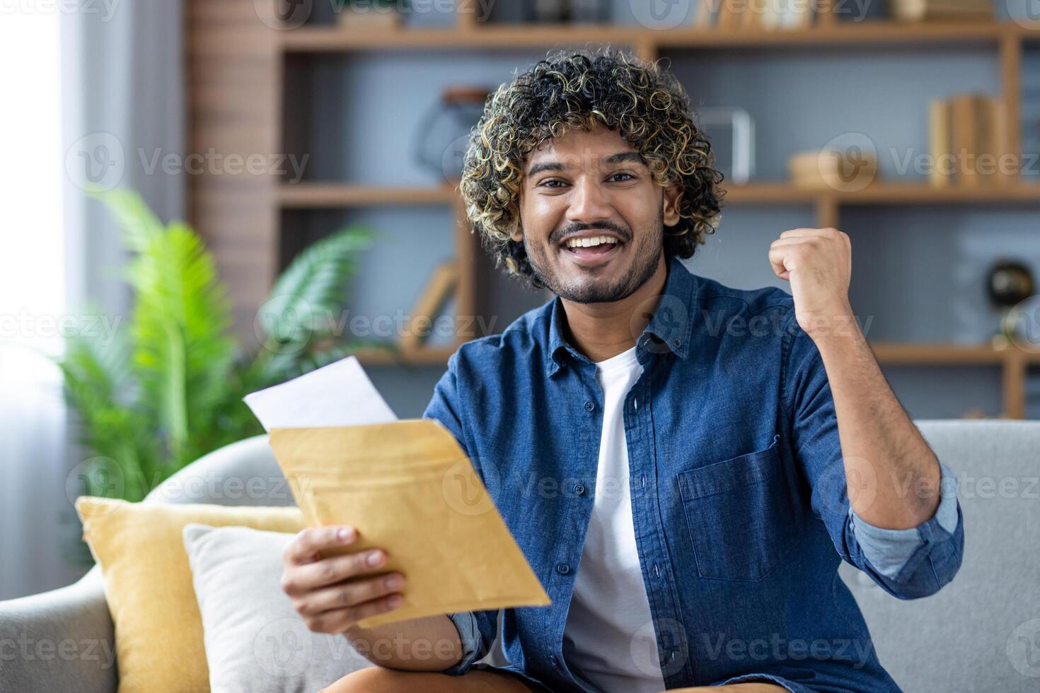 Winning good news, man with mail envelope looking joyfully at camera, sitting in living room at home, received notification message by mail about good results photo