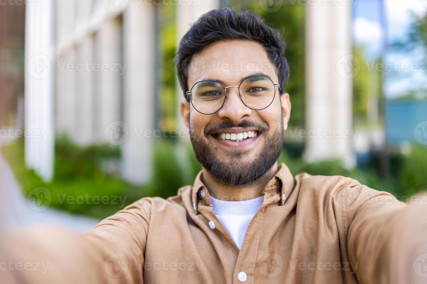 Close-up of smiling man taking selfie outdoors on sunny day. Man with beard and glasses in casual clothing enjoying outdoor moment. Concept of happiness, modern communication, and casual lifestyle. photo