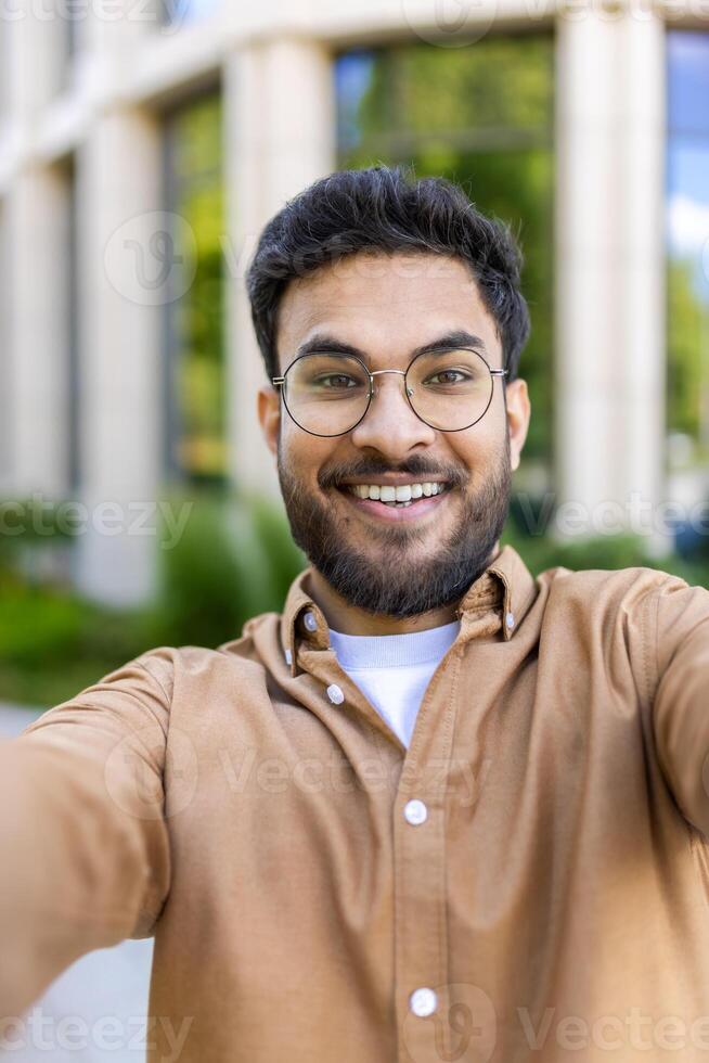 Cheerful hispanic man taking selfie outside office building on sunny day photo