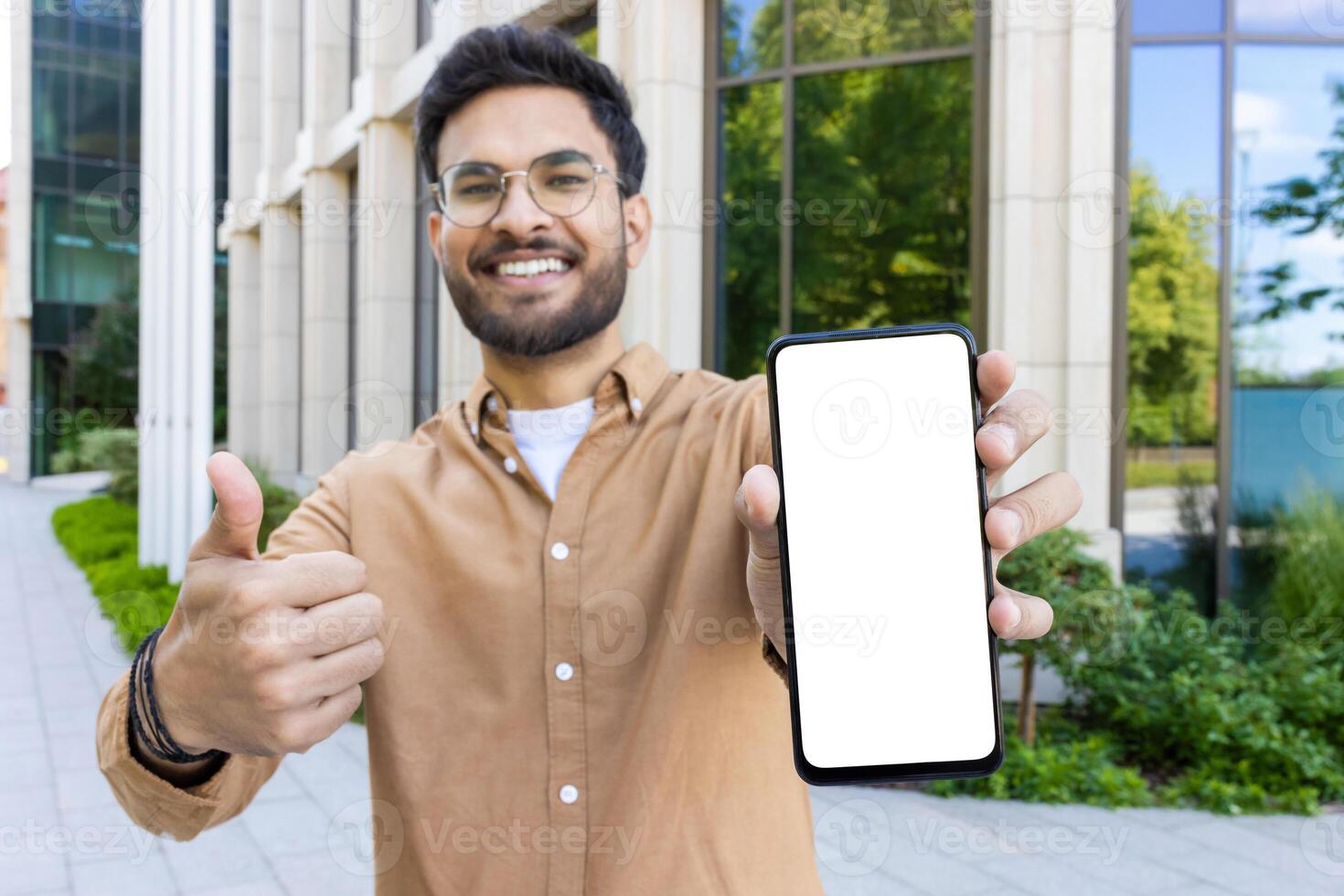 Smiling man showing blank smartphone screen with thumbs up outside modern building photo