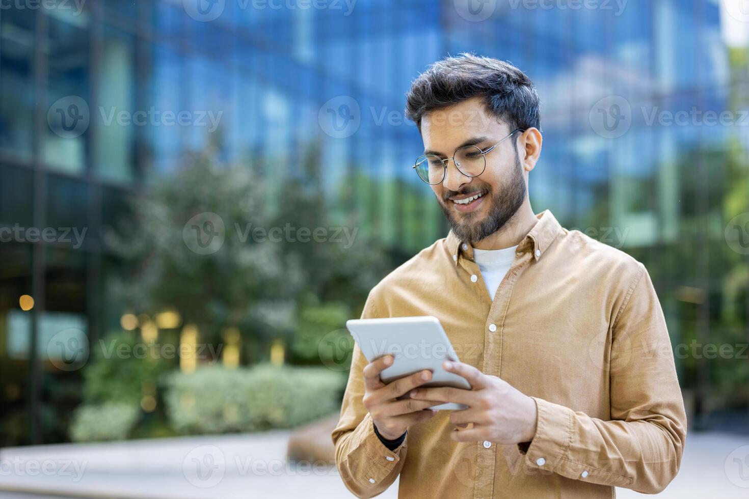 Young man using tablet outside modern office building, smiling and reading photo