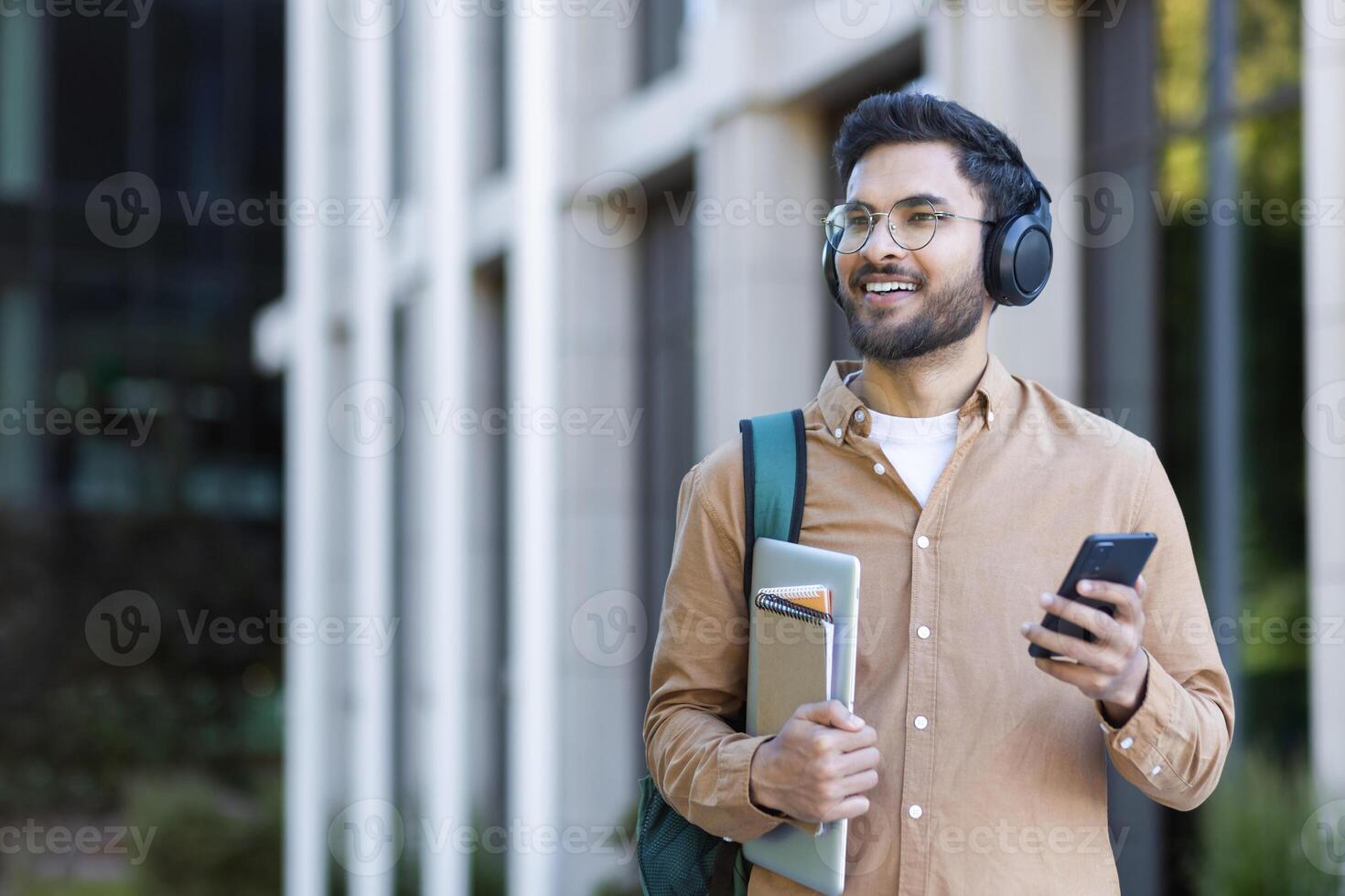 Smiling student wearing headphones holding laptop and phone outdoors in urban setting with confidence and style photo
