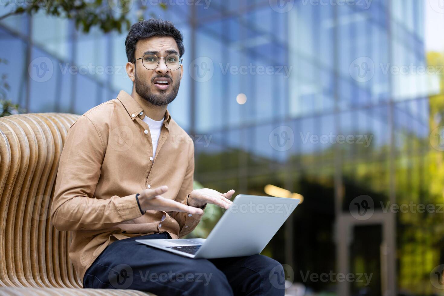 Confused man using laptop outdoors with modern glass building background, expressing frustration or misunderstanding during work, business or study session photo
