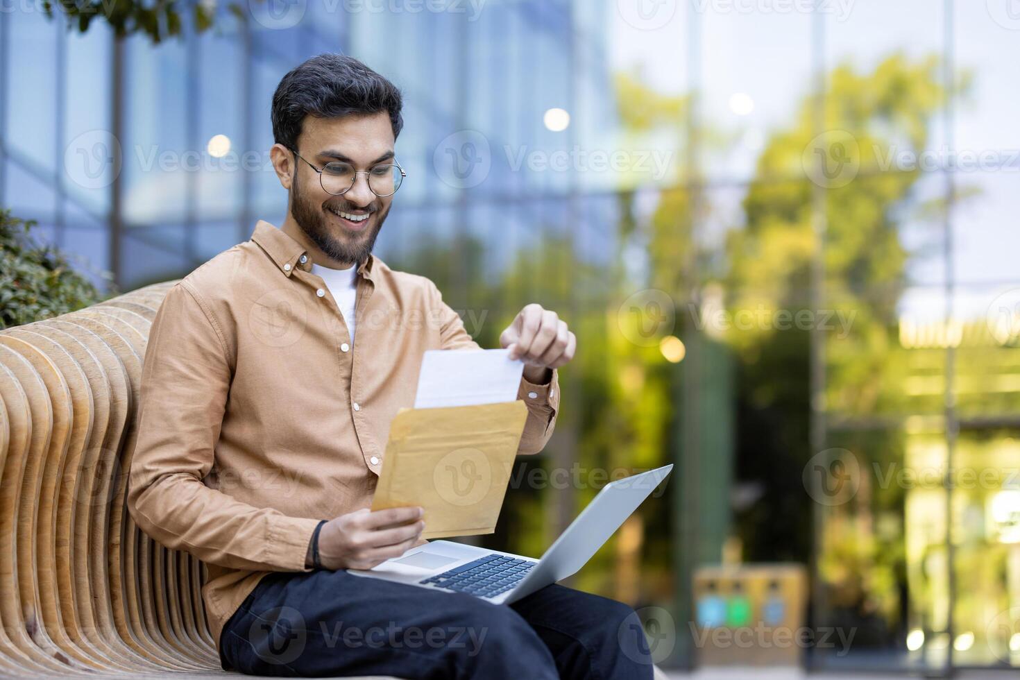 Man reading letter outdoors while sitting on wooden bench with laptop in urban setting photo