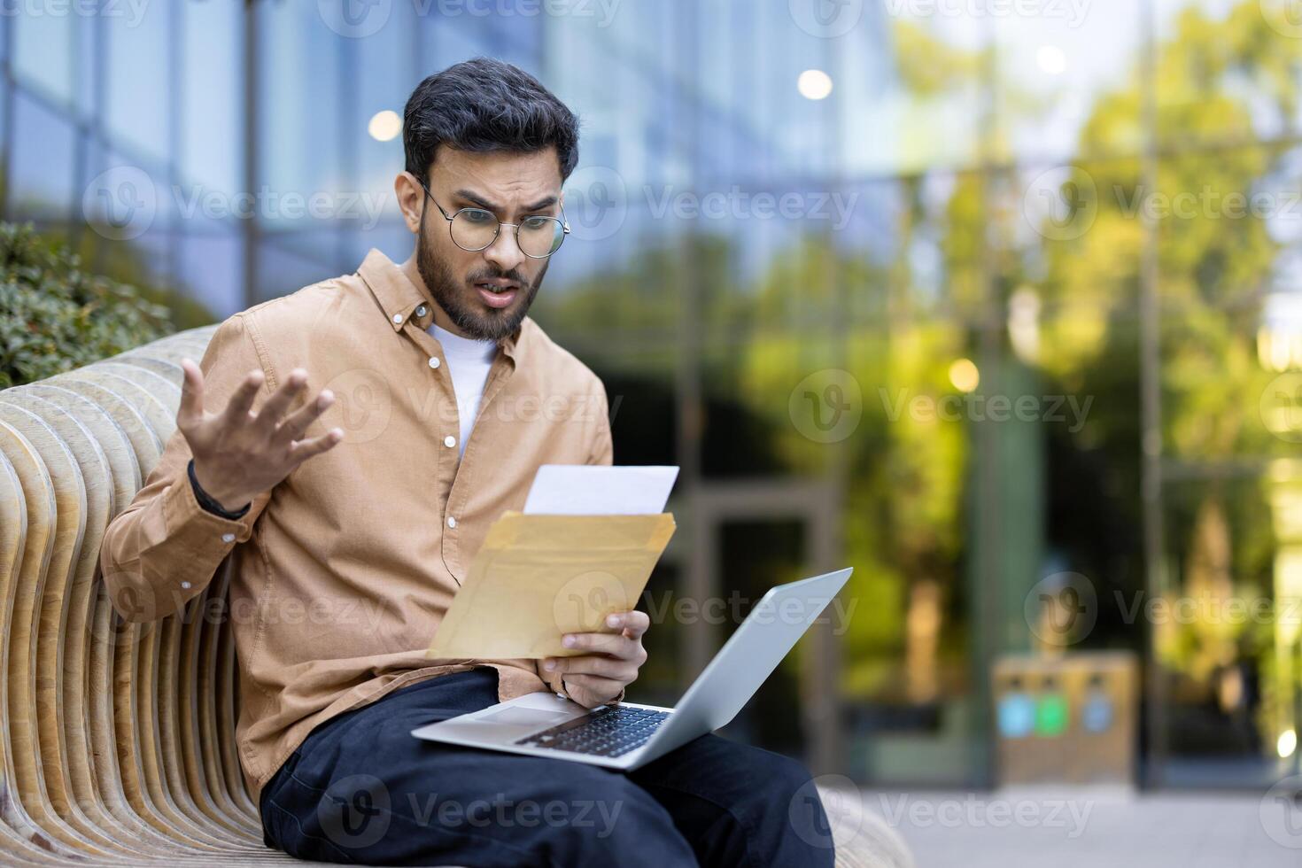 Shocked young man reading unexpected news in letter while sitting outside with laptop open, expressing surprise and confusion photo