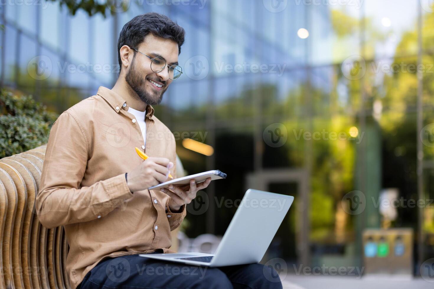 Smiling young professional takes notes on tablet while engaged in online learning with laptop in modern outdoor workspace photo