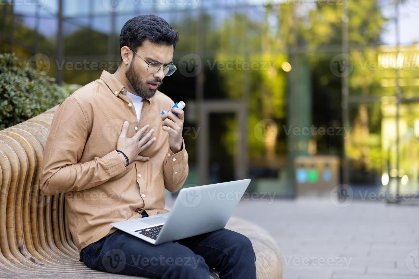 Man experiencing chest discomfort while using an inhaler outdoors with laptop on bench photo