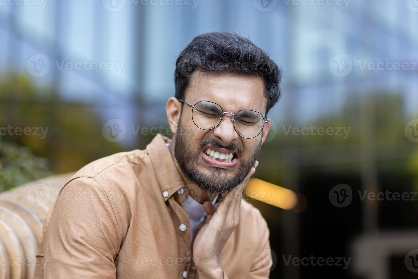 Man with glasses experiencing severe toothache outside office building, showing discomfort while holding cheek photo