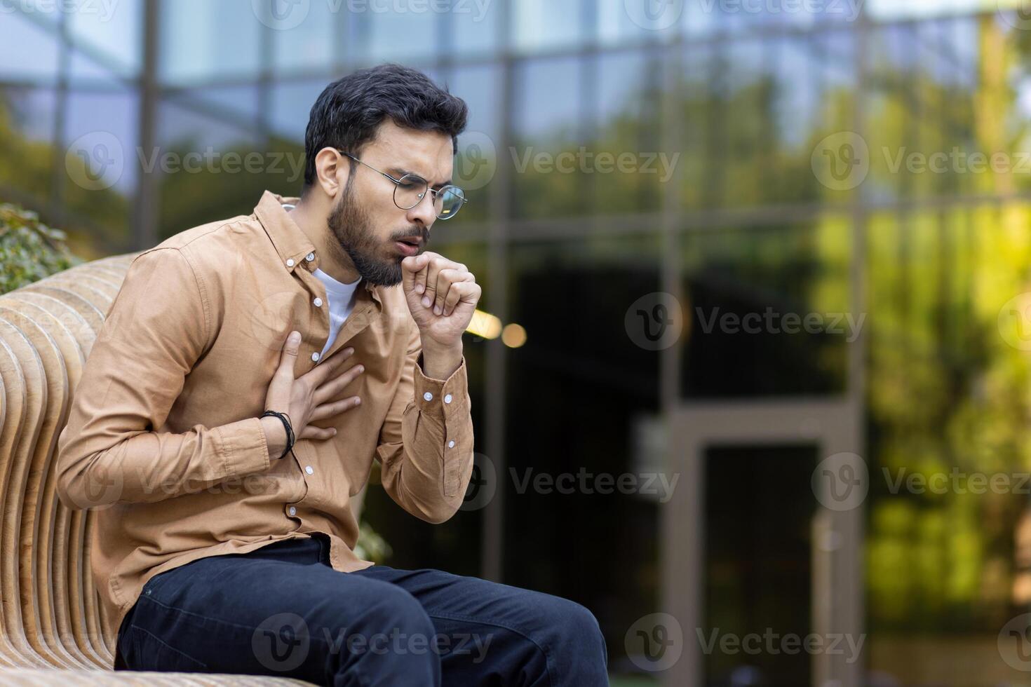 Young man experiencing discomfort and coughing while seated outdoors, highlighting health issues, allergies, or respiratory concerns in an urban environment photo