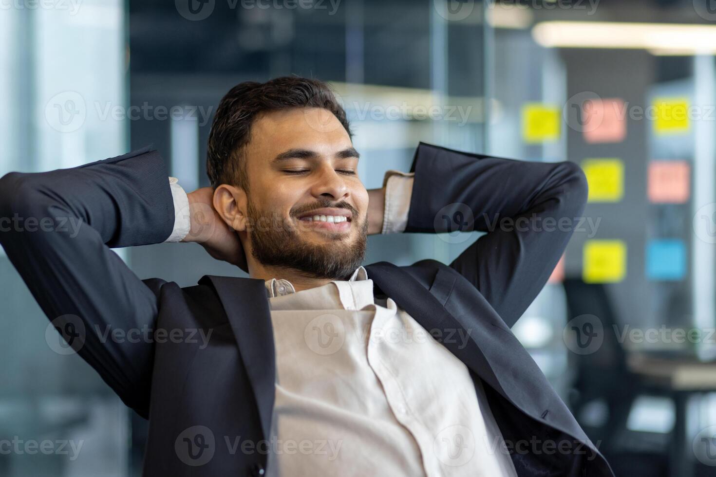 A well done man rests with his hands behind his head, dreams and thinks about the future results of his achievement. A businessman at his workplace inside the office works close-up in a business suit photo
