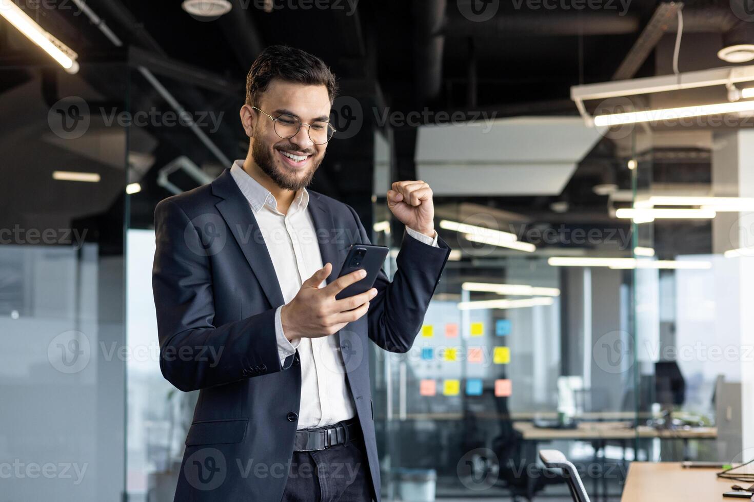 A businessman in a suit celebrates success in a modern office while looking at his smartphone, showcasing excitement and achievement in a professional setting. Bright office lights emphasize his joy. photo