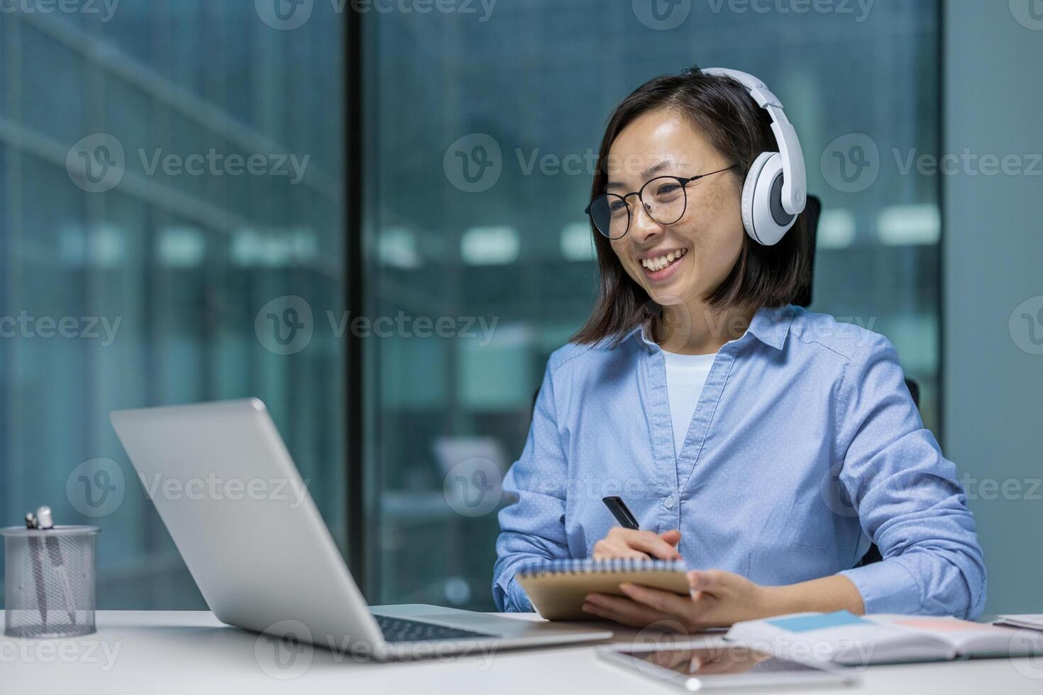 Woman watching online course, training conference inside office at workplace. Asian Office worker in headphones using laptop for remote meeting, writing data in notebook photo