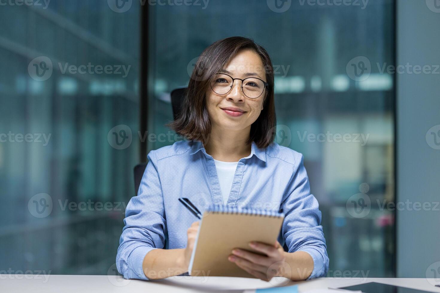 An Asian woman in glasses sits at a desk holding a notebook, looking directly at the camera with a warm, friendly smile photo