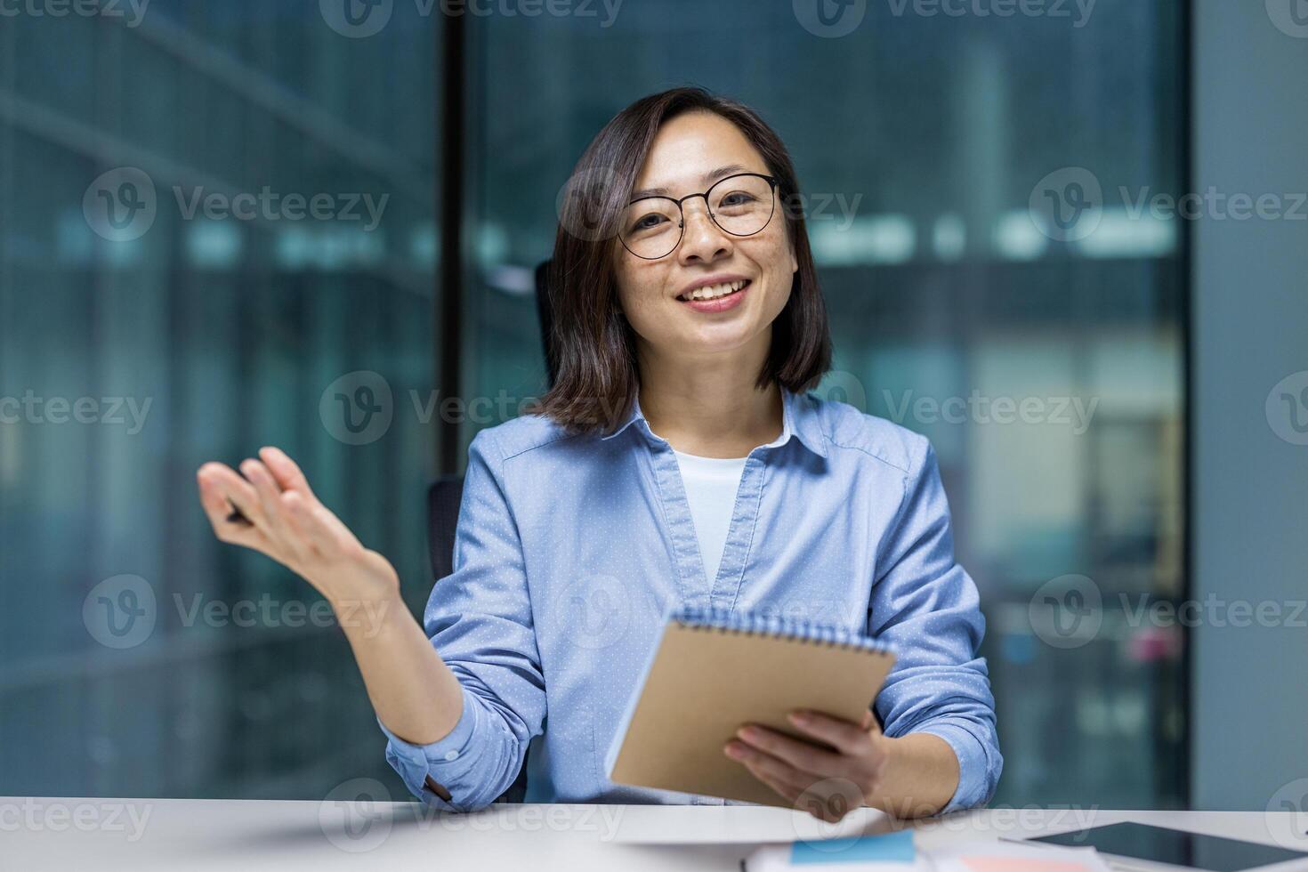 Webcam view, woman holding a notebook in motion, smiling and looking at the camera. Asian woman on online business meeting conference, recording data and studying inside office at workplace. photo