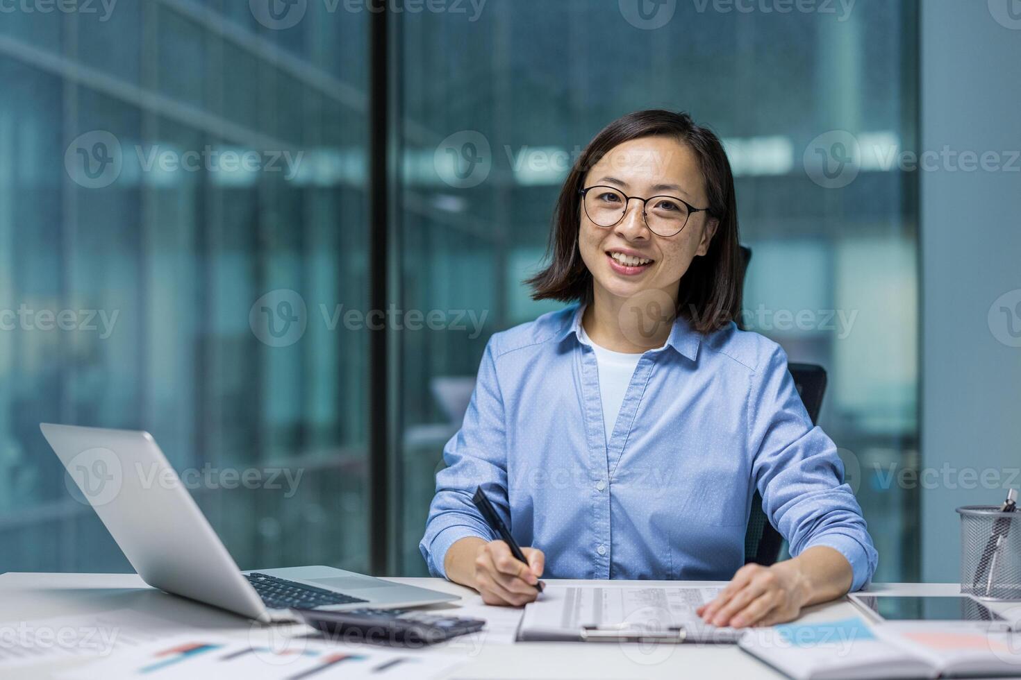 A young Asian woman is working at her office desk, with a laptop and financial documents, smiling at the camera. photo