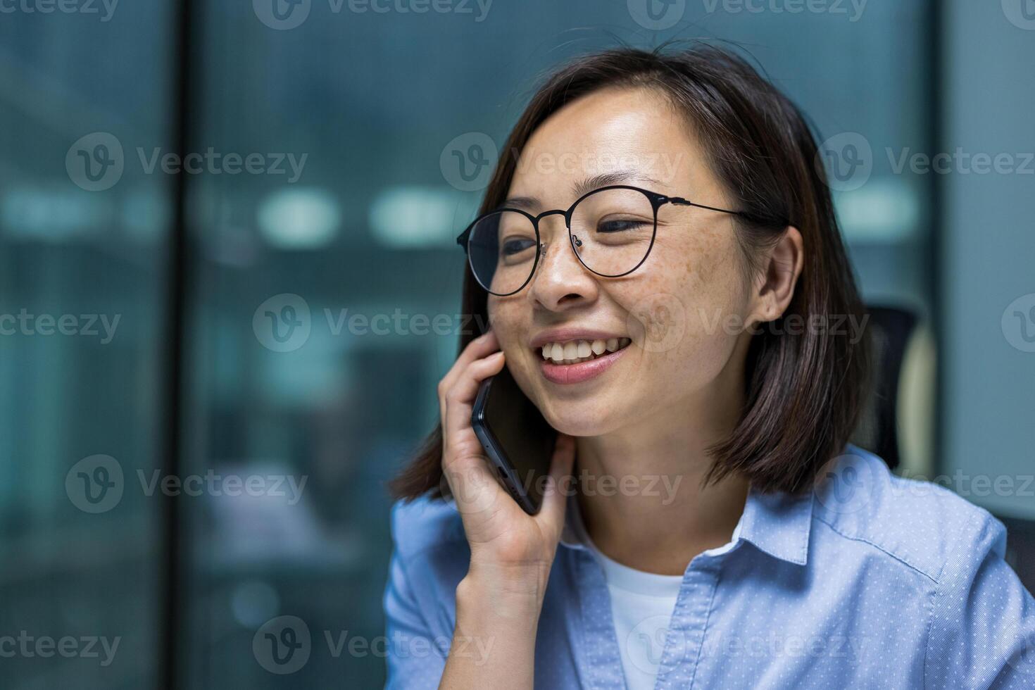 Young successful businesswoman talking on the phone inside the office close up. Satisfied Asian woman smiling, working near the window. photo