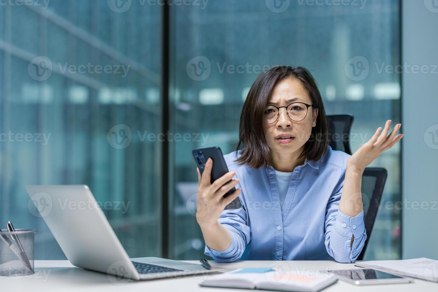 Frustrated asian businesswoman at her desk looking at her phone with a confused expression in a modern office setting. photo