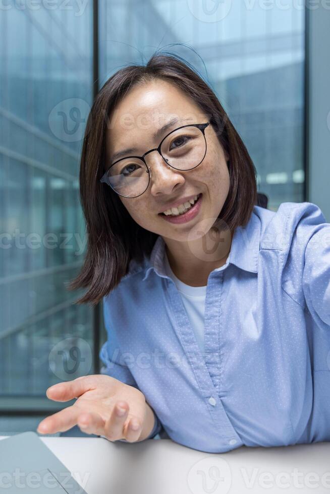 Young businesswoman using phone app for call, smartphone camera view. Office worker looking at camera smiling and talking to colleagues and partners sitting at desk inside office. photo