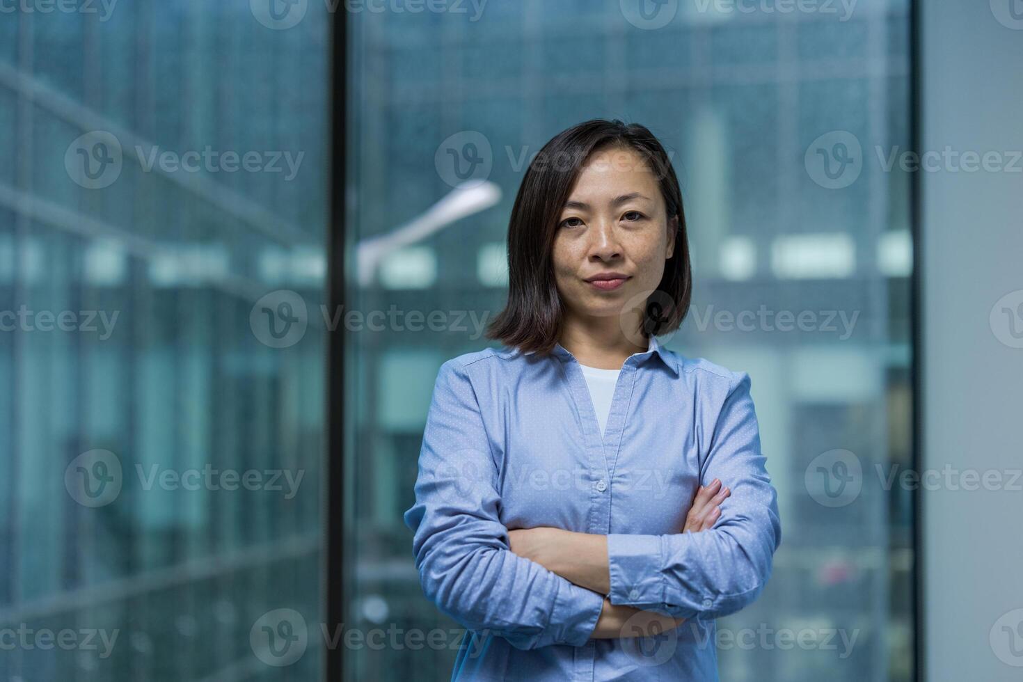 Portrait of an Asian businesswoman standing confidently with her arms crossed against the backdrop of a modern office building window. photo