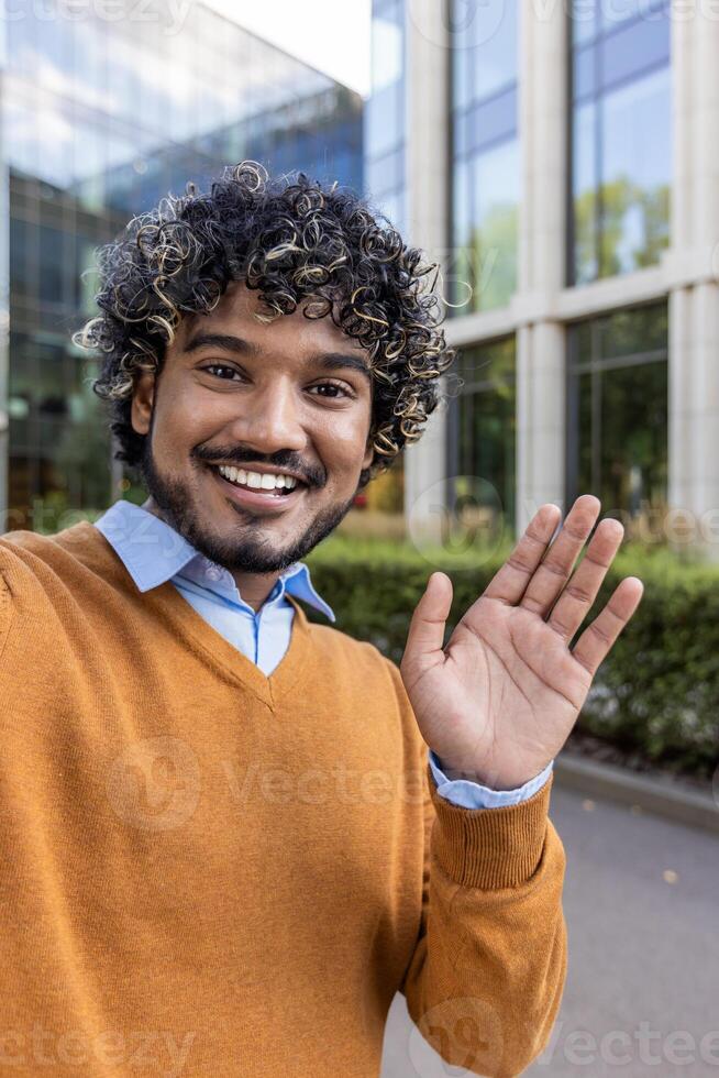Smiling businessman in casual attire waving at camera during call using phone. Image captures friendly, cheerful mood in urban business setting, with modern office in background. photo