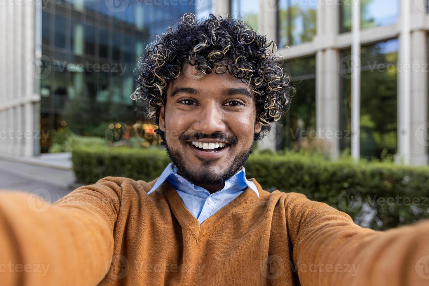 Smiling young man taking selfie outside modern office building, wearing casual attire. Capturing cheerful moment with phone for call. Bright outdoor setting in urban environment photo