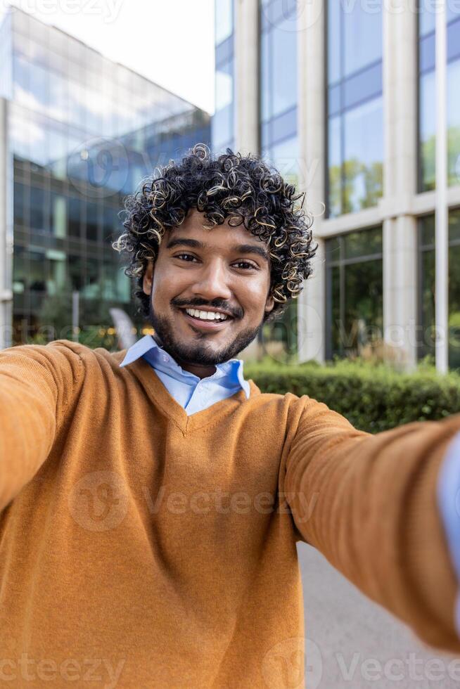 Confident young man with curly hair captures a selfie using phone outside contemporary office building expressing positivity. Reflects modern lifestyle, city life, self-expression, and technology. photo