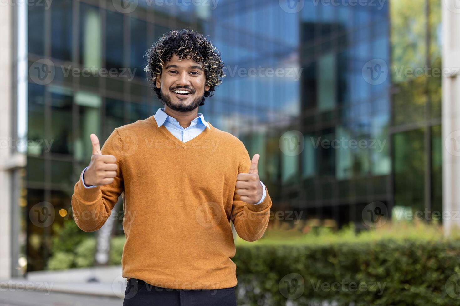 Confident young professional showing positivity approval outside corporate office, wearing casual orange sweater. Represents success, motivation, career growth, job satisfaction, positive attitude photo