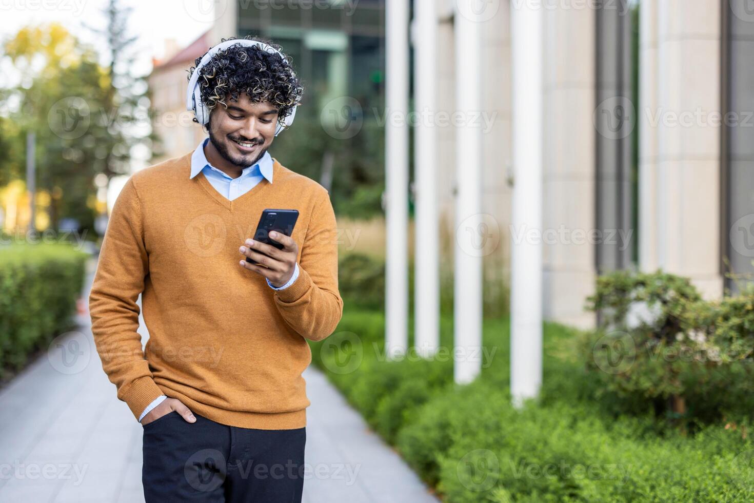 Young man with headphones using smartphone while walking on street. Smiling in casual clothes, enjoying music and technology outside in urban environment. Represents modern communication photo