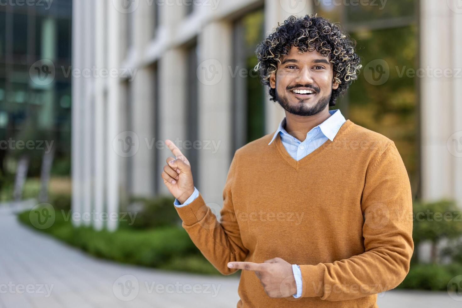 Smiling young professional stands outside modern office building, confidently pointing with both hands. Image conveys positivity, success, and enthusiasm in business or academic environment. photo