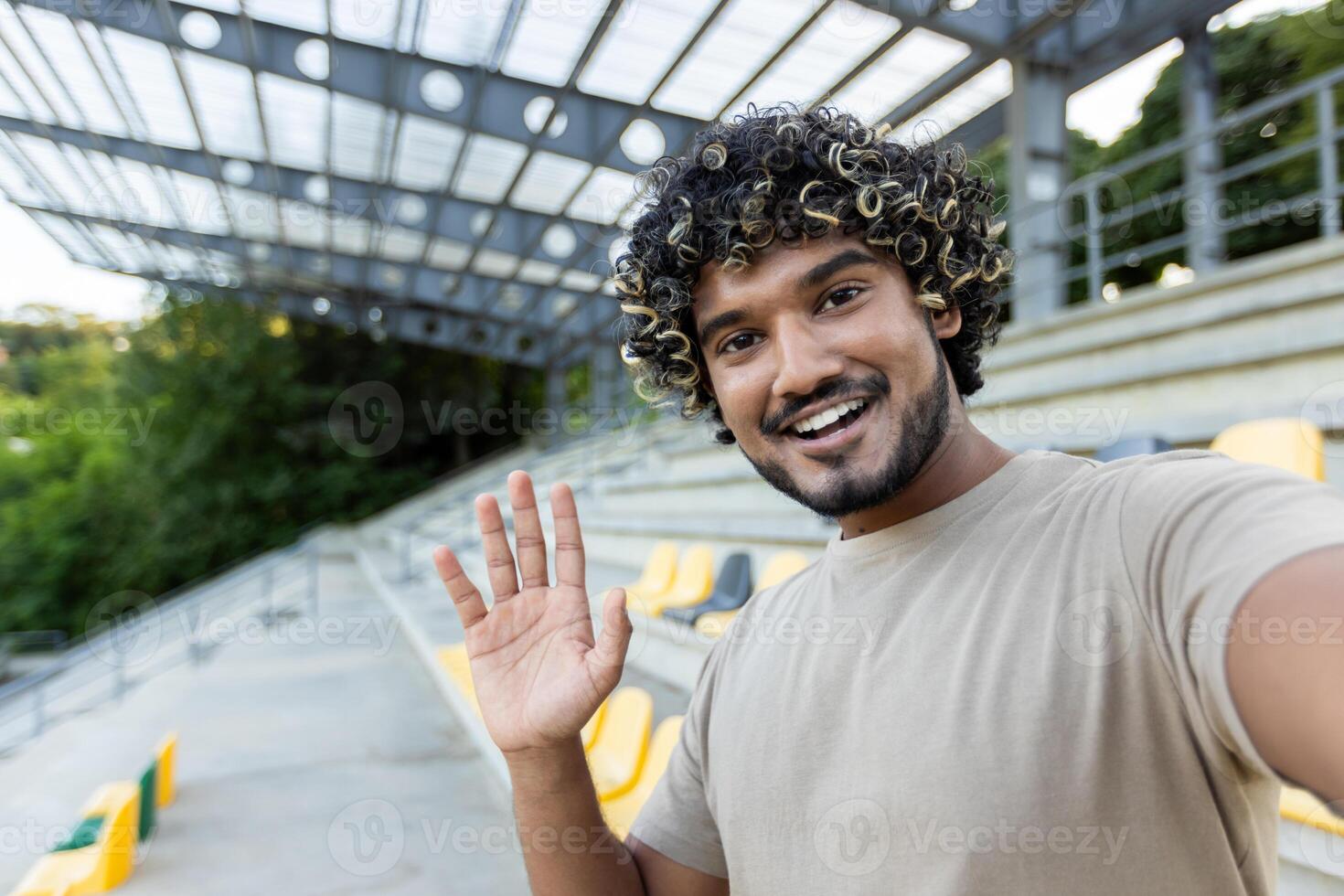 A smiling man with curly hair waves during a call, sitting in the stands of an empty stadium, looking directly at the camera photo