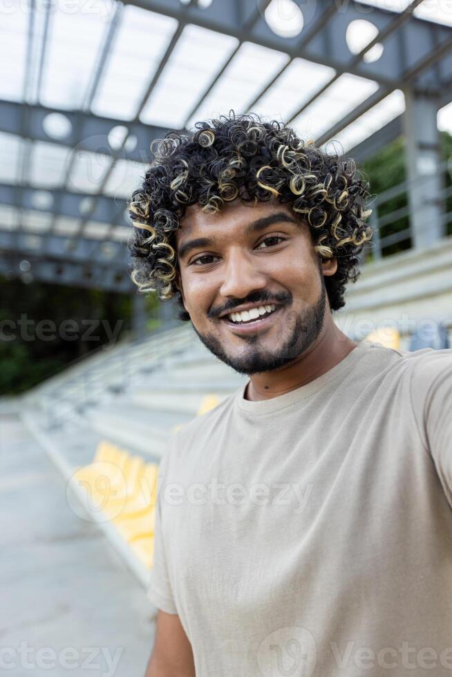 A young man smiles into the camera while calling at the stadium, in a casual setting with seats visible in the background photo