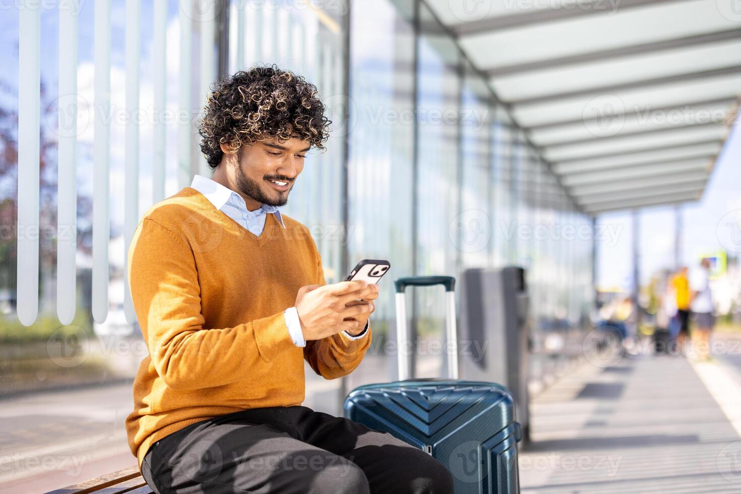 A young businessman with curly hair using his mobile phone while waiting at the bus station with his luggage photo