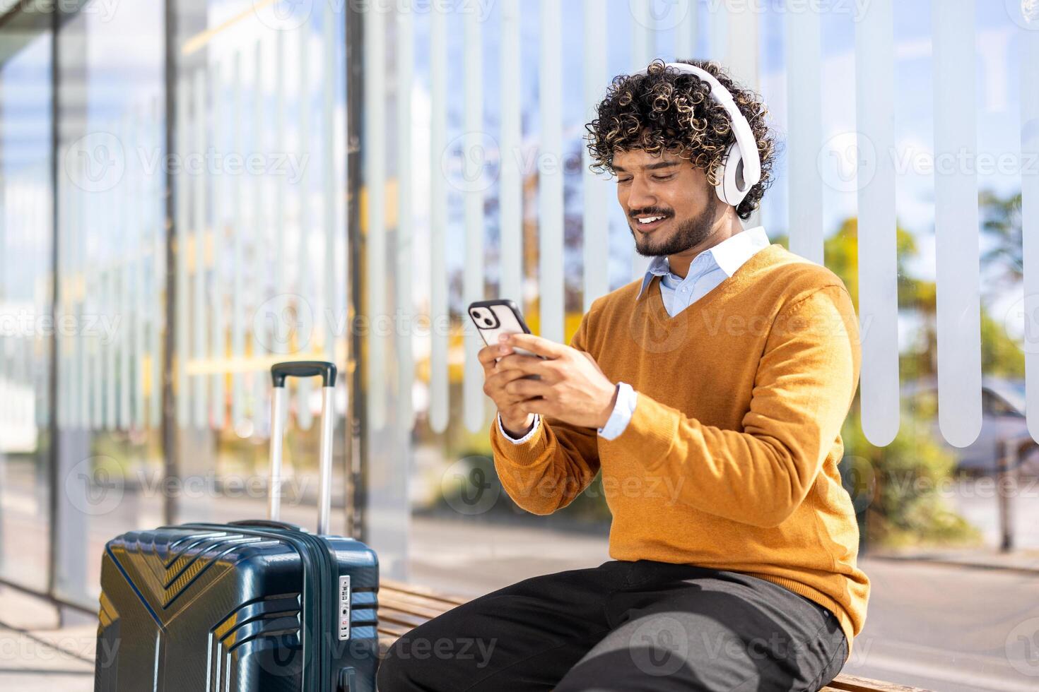 A young man with headphones waits at a bus stop, looking at his smartphone with his suitcase beside him on a bright day. photo