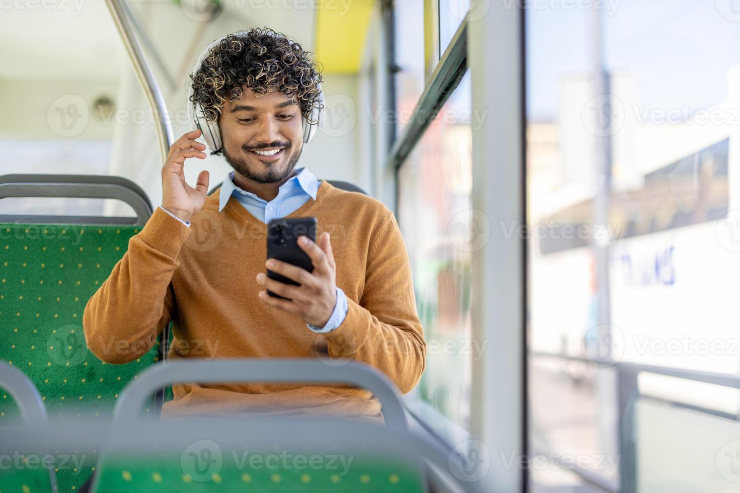 A curly-haired man enjoys music on headphones and checks his phone while riding public transportation in a city. photo
