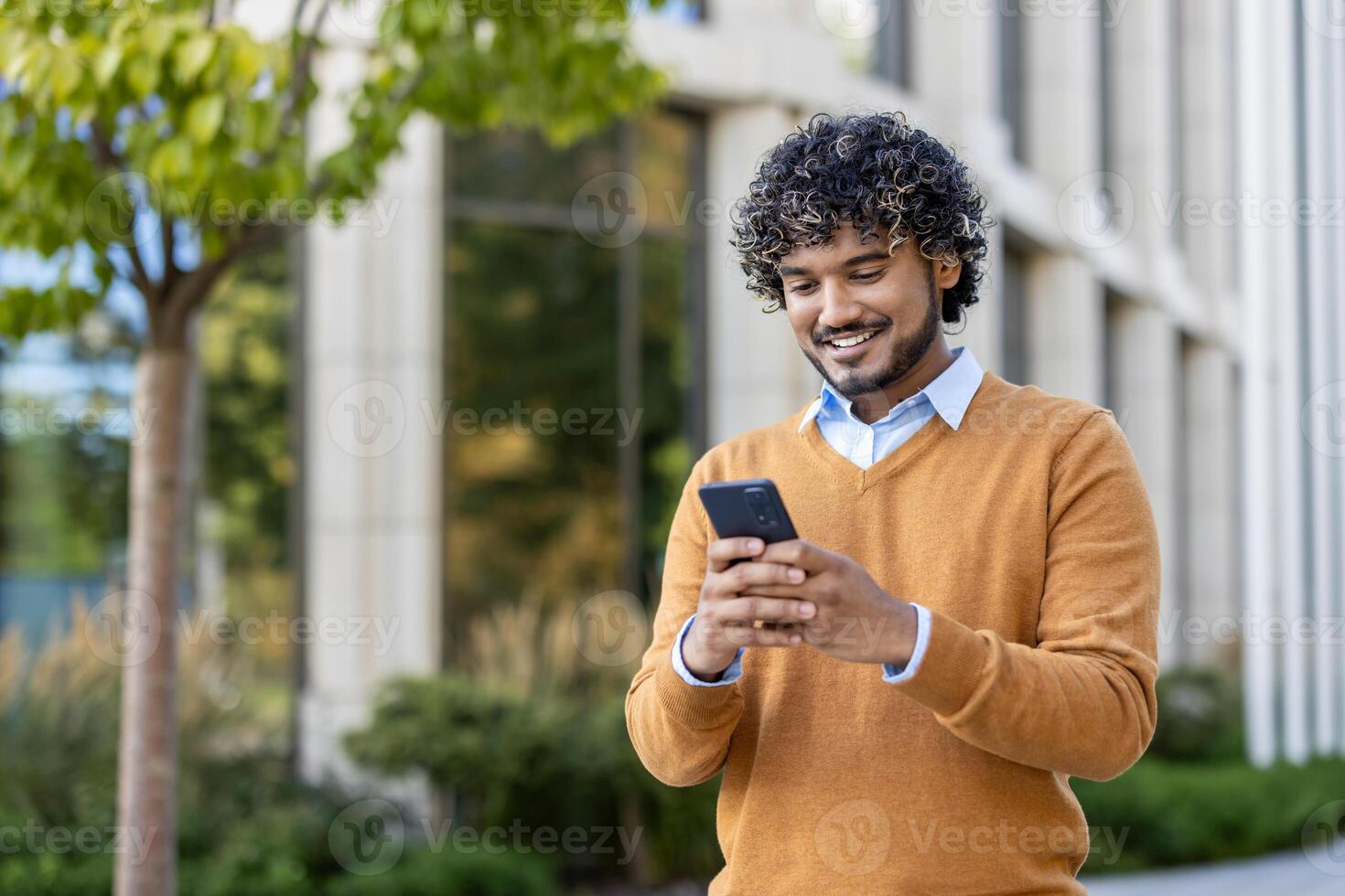 Young professional with curly hair using smartphone outdoors. Scene conveys connectivity, technology, and communication. Individual dressed in casual autumn attire, standing confidently near office photo