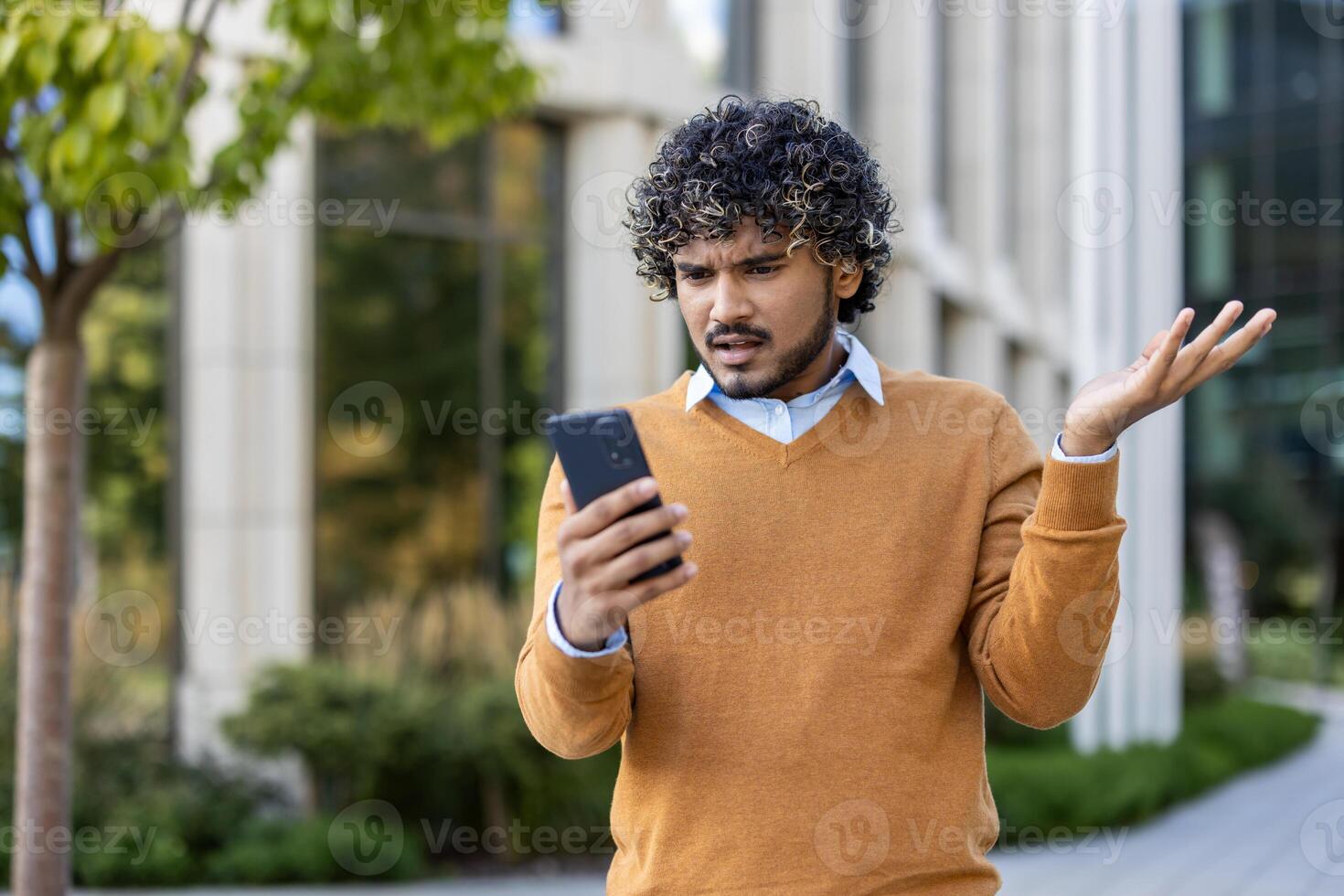 Young man appears perplexed as he looks at smartphone, expressing confusion and frustration. Dressed in casual attire with orange sweater, he stands outside in urban environment photo