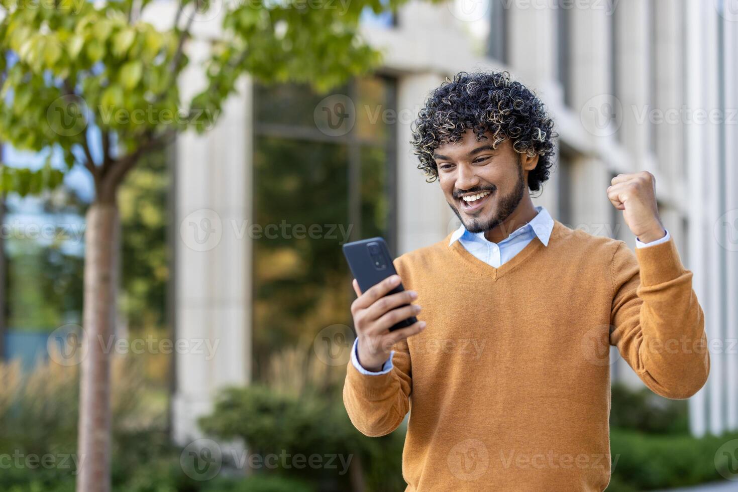 Young man in casual attire celebrating success while looking at smartphone outdoors. Excitement and triumph visible as he raises fist in joy, embodying achievement and happiness. photo