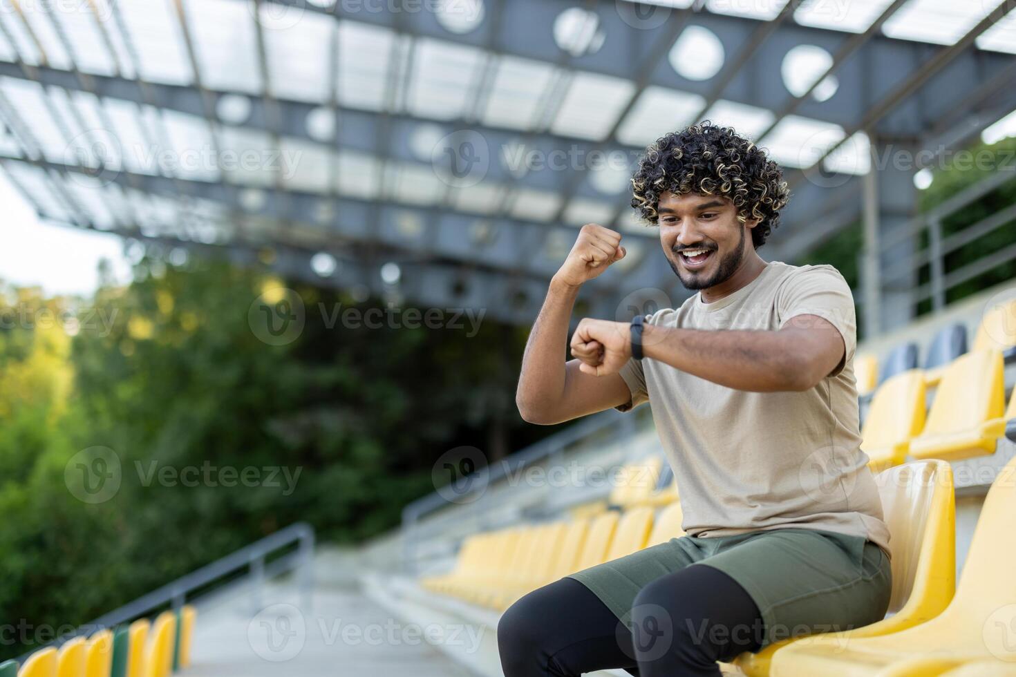 Young athlete celebrates successful training results, resting while sitting. Man checks data on smart watch fitness bracelet, smiles contentedly, holds hand up. photo